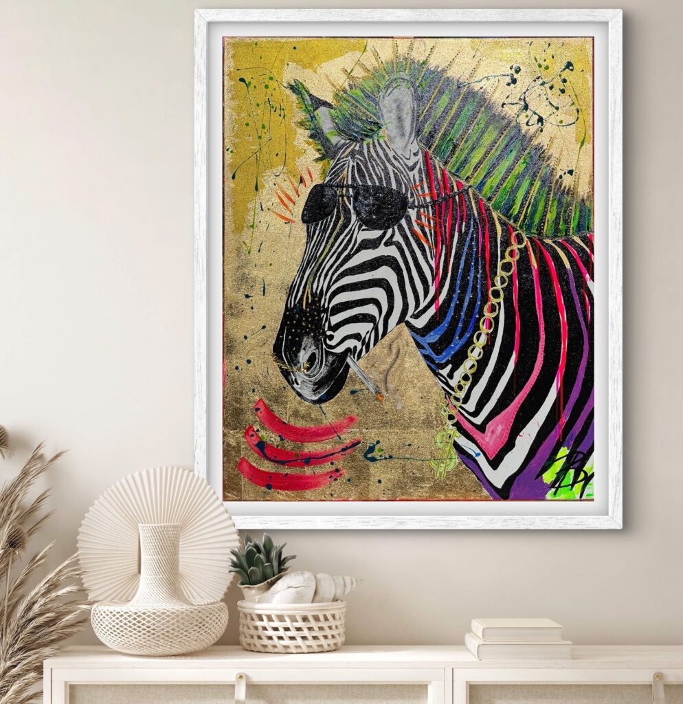 A colorful artistic depiction of a zebra wearing sunglasses and a chain necklace, with a vibrant, punk-style mane. The background is a metallic gold with splashes of paint in various colors. The artwork is framed in white and displayed on a shelf with decorative items.