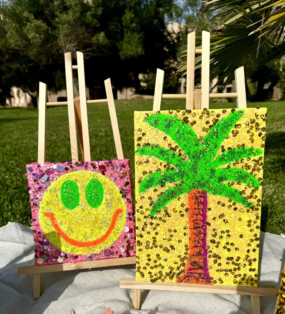 Two colorful art canvases on wooden easels are displayed outdoors on a grassy surface. The left canvas features a bright yellow smiley face with green eyes on a pink background, adorned with sequins. The right canvas showcases a palm tree design with a brown trunk and green leaves on a yellow background, also embellished with sequins and glitter.