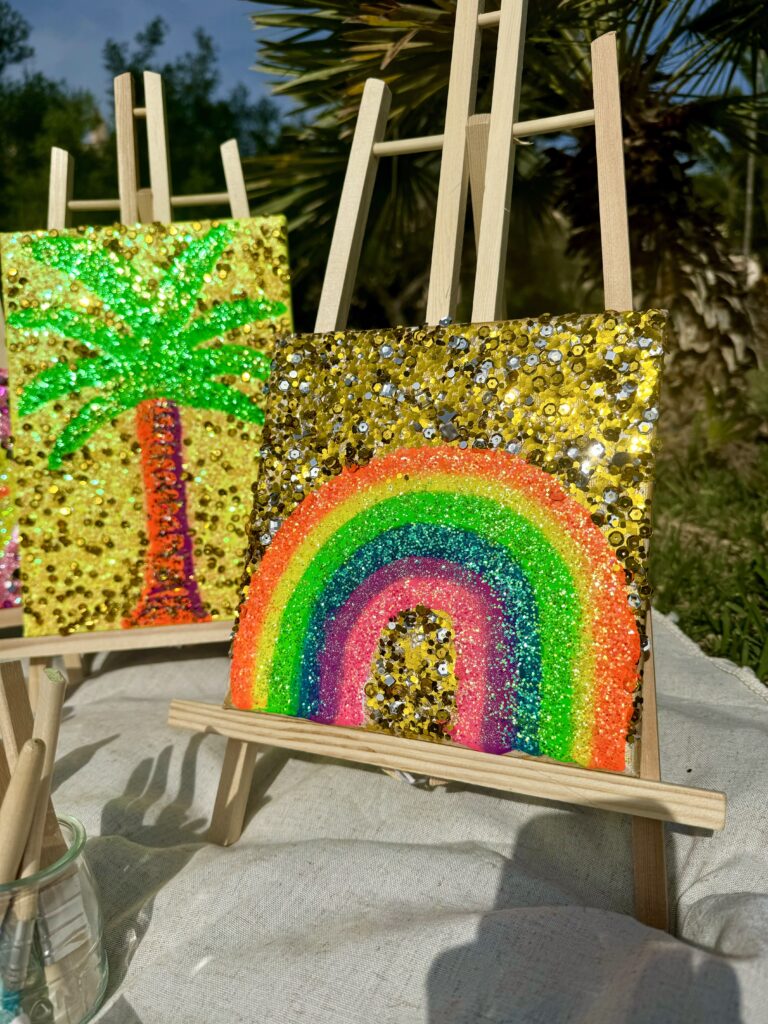 Two colorful, glittery artworks are displayed on small easels. One artwork features a palm tree in vibrant colors against a golden background, while the other depicts a rainbow with bright arcs surrounded by sparkling sequins. The setting includes green foliage in the background and a cloth surface beneath the easels.