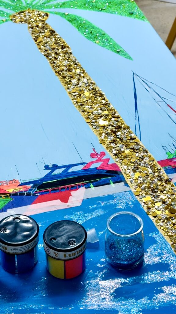 A colorful art piece featuring a blue background with a golden glittery stripe. The design includes stylized boats in vibrant colors near the bottom, and embellishments of green palm leaves at the top. In the foreground, there are several containers of glitter and paint, including a blue glitter jar and three paint pots in blue, yellow, and pink.