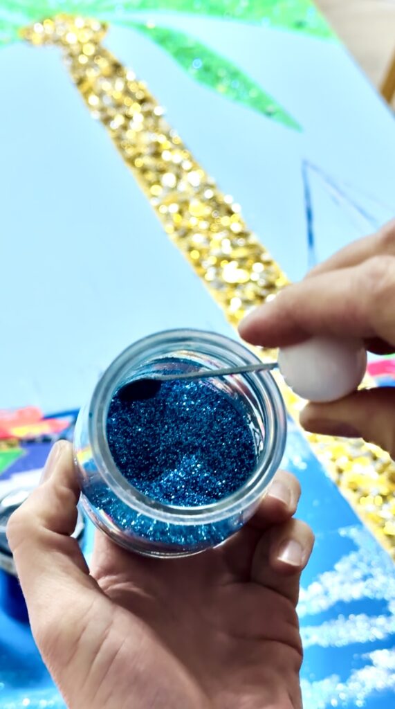 A hand holds a jar of blue glitter with a small tool poised to scoop some out. In the background, a colorful art piece featuring a palm tree and golden accents is partially visible.