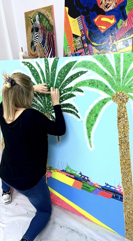 A person is applying glitter to palm tree designs on a colorful wall mural. The background is a bright blue, and there's a colorful depiction of boats at the bottom. Other artworks, including a zebra and a superhero, are visible in the background.