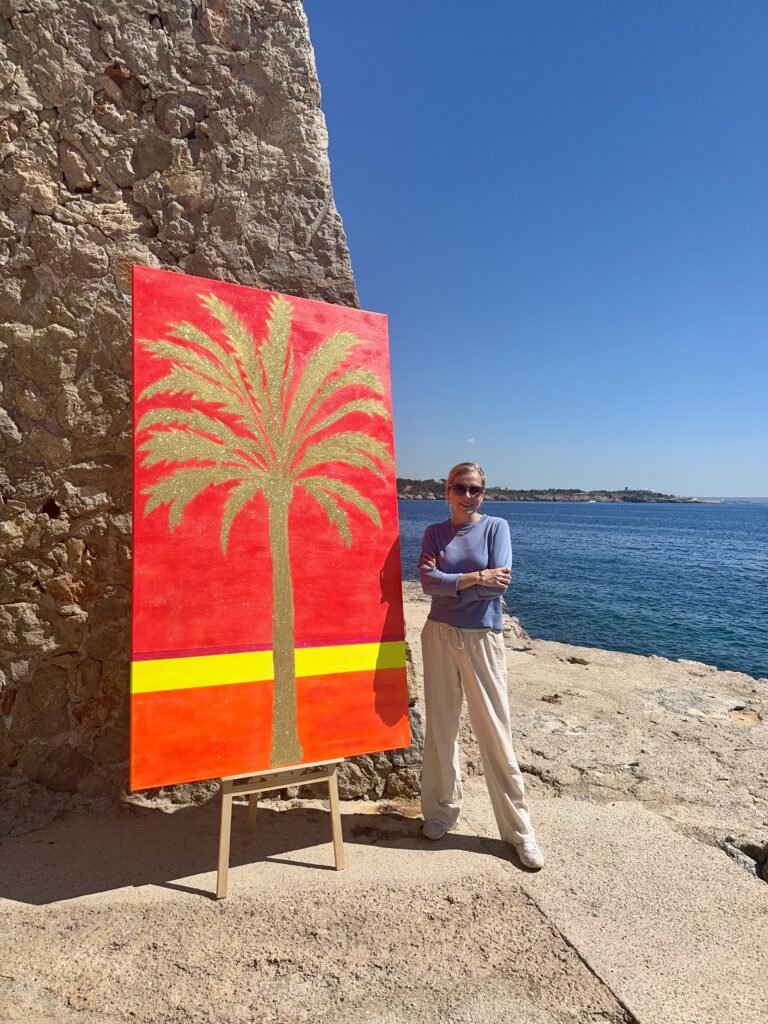 A person stands next to a large painting of a palm tree, set against a bright red background, on a rocky shoreline by the sea. The sky is clear and blue, and the person is wearing a light blue top and beige trousers.