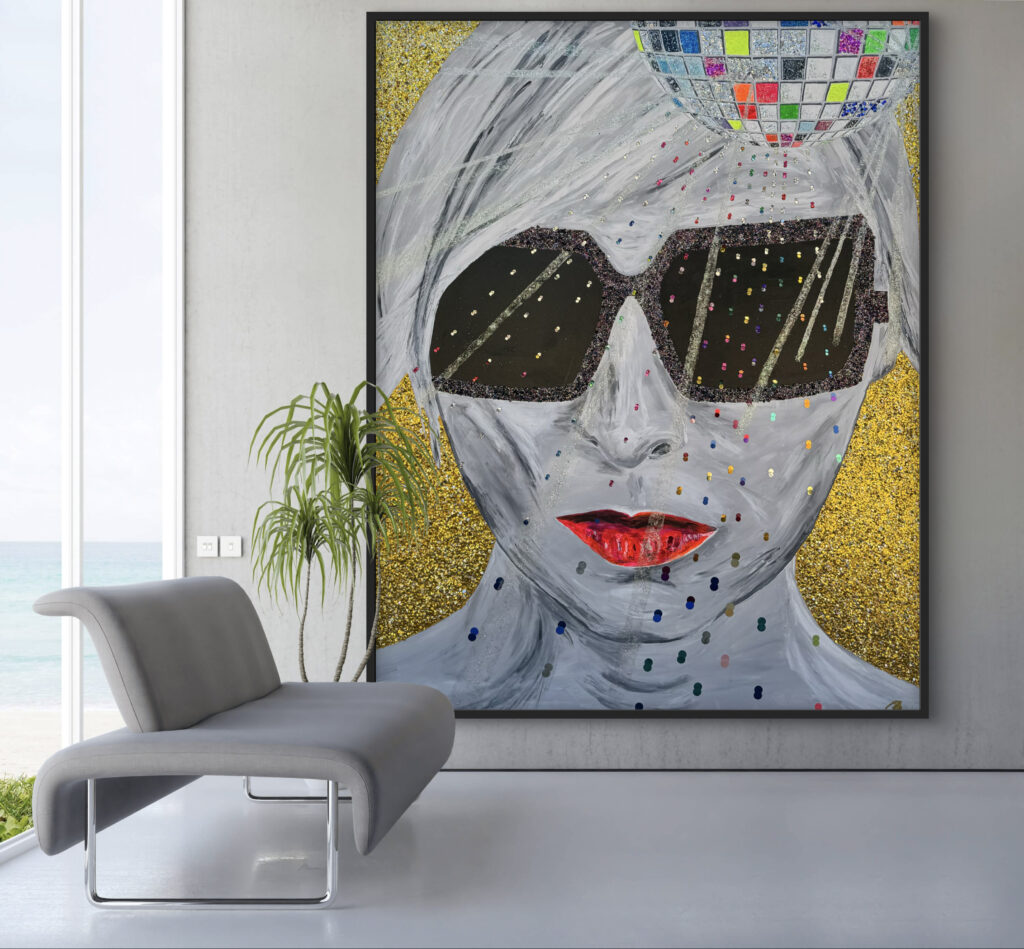 A modern interior features a gray chair in front of a large, colorful artwork. The painting depicts a stylized figure with a pale face, wearing dark sunglasses and bright red lips. The background is adorned with gold glitter and colorful dots, while a disco ball is illustrated above the figure's head. Natural light streams in through large windows, revealing a serene view of the ocean.