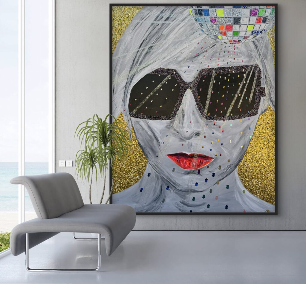 A modern interior features a gray chaise lounge beside a large artwork depicting a stylized face with white hair, wearing dark sunglasses and bright red lips. The background is gold and adorned with colorful dots, while the space is filled with natural light from large windows showcasing an ocean view.