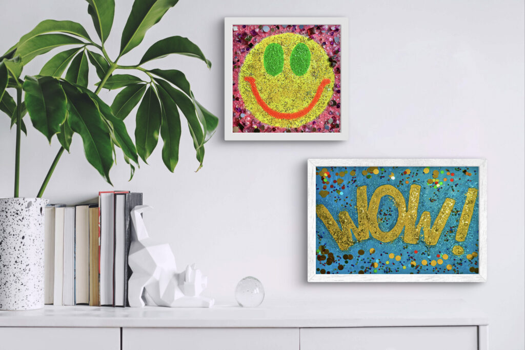 Alt text: A modern interior scene featuring a large green plant in a speckled white pot, two framed artworks on the wall—one with a smiling face in yellow and green on a pink background, and the other displaying the word "WOW!" in gold on a blue background. A stack of books and a white, geometric cat sculpture are visible on a shelf below.