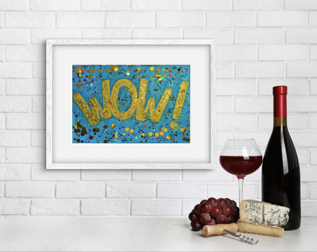 A framed artwork featuring the word "WOW!" in gold glitter against a blue splattered background is displayed on a table. Next to it, there is a glass of red wine, a bottle of red wine, and an assortment of cheeses and grapes. The backdrop consists of a white brick wall.