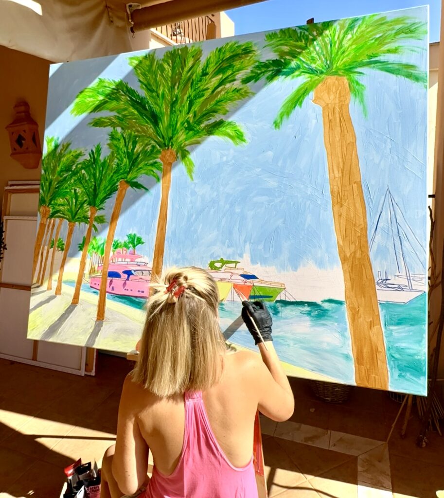 A woman with blonde hair, wearing a pink tank top, paints palm trees and boats on a large canvas outdoors. The scene features vibrant colors with a blue sky and water, depicting a sunny day. The artist holds a paintbrush and is focused on her work.